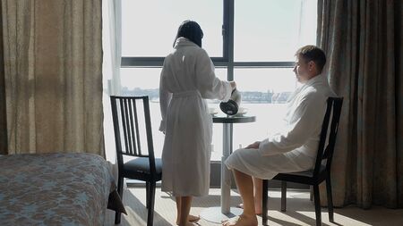 Young happy couple sitting by the window in hotel, at home. A woman and a man drink tea or coffee. A familyの写真素材