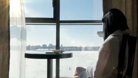 A young brunette woman sits at a table by the window and drinks tea, coffee. Morning, bright sunの写真素材