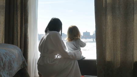 Mother and her daughter are sitting by the window. A woman and a child are looking out the window. Sunny morningの写真素材