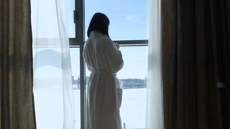 Young beautiful girl standing by the big window talking on the phone. A woman in a white robe is standing by the windowの写真素材