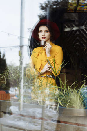girl in a cafe with a cup of coffee and a hat. beautiful girl with a cup of coffee near the window in a cafe, waiting for a meeting, breakfastの写真素材