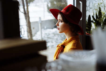 girl in a dress and a hat looks out the window, sad, self-isolation, depression, pandemic, virus