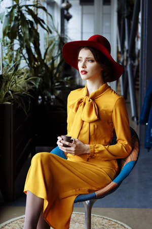 girl in a cafe with a cup of coffee and a hat. beautiful girl with a cup of coffee at the window in a cafe, waiting for a meeting, breakfastの写真素材