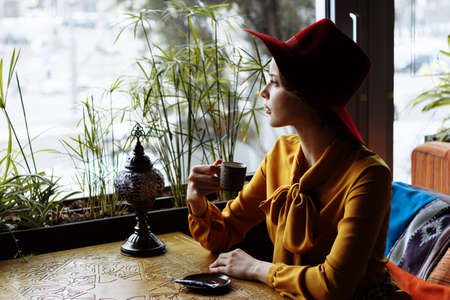 girl in a cafe with a cup of coffee and a hat.portrait of sensual young girl wearing floppy hat and blouse with bow. Beautiful brunette woman in cafe holding cup of coffeeの写真素材