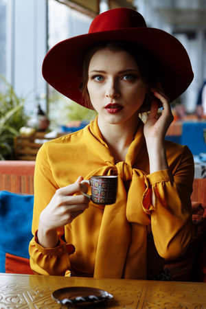 girl in a cafe with a cup of coffee and a hat.portrait of sensual young girl wearing floppy hat and blouse with bow. Beautiful brunette woman in cafe holding cup of coffeeの写真素材