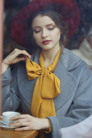 girl in a cafe with a cup of coffee and a hat. beautiful girl with a cup of coffee at the window in a cafe, waiting for a meeting, breakfast watch through the glass of a girl sitting in a cafeの写真素材