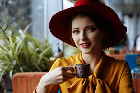 girl in a cafe with a cup of coffee and a hat. beautiful girl with a cup of coffee at the window in a cafe, waiting for a meeting, breakfastの写真素材