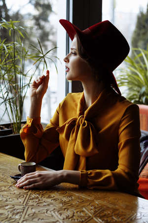 girl in a cafe with a cup of coffee and a hat. beautiful girl with a cup of coffee at the window in a cafe, waiting for a meeting, breakfastの写真素材