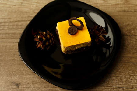 chocolate cake with yellow icing and cream lies on a black plate, next to it there are two lumps, decor, on a wooden tableの写真素材