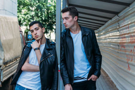 beautiful and attractive guy is standing in the city, posing. dressed in a black leather, white T-shirt, jeans and white shoes. walk around the city in black glasses. communication. meeting friends.の写真素材