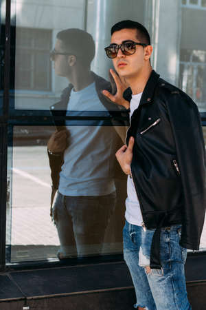 young, handsome guy in a white T-shirt, black, leather jacket and blue, torn jeans is on the street, model, city walk, live style. in white sneakersの写真素材