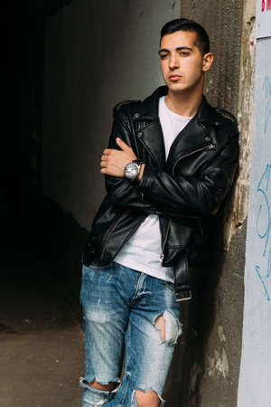 young, handsome guy in a white T-shirt, black, leather jacket and blue, torn jeans is on the street, model, city walk, live style. in white sneakersの写真素材