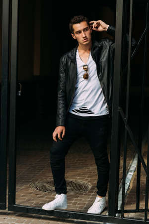 young, handsome guy in a white T-shirt, black, leather jacket and black pants is on the street, n...