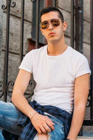 Attractive, sexy guy sits leaning on a curb on a ledge in the wall on the street in sunglasses. self-confidence, flatulence and arrogance.model, posing, portrait of a young manの写真素材