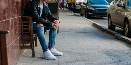 attractive guy sitting on a bench in a cafe on the street, white sneakers, white T-shirt, black leather jacket and black jeans, pants. model. waiting for a meeting.の写真素材