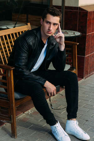 attractive guy sitting on a bench in a cafe, holds sunglasses in his hand, on the street, white sneakers, white T-shirt, black leather jacket and black jeans, pants. model. waiting for a meeting.の写真素材