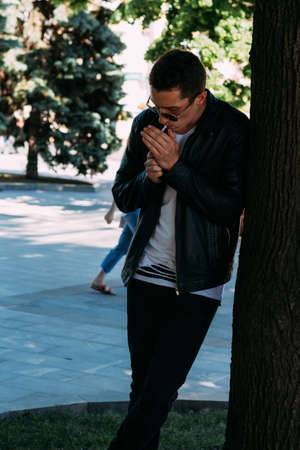 a young guy smokes near a tree in the city. A black jacket, jeans, white sneakers, a white T-shirt and glasses. bad habit. smoking in a public placeの写真素材