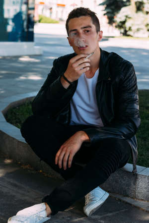 a young guy smokes near a tree in the city. A black jacket, jeans, white sneakers, a white T-shirt and glasses. bad habit. smoking in a public placeの写真素材