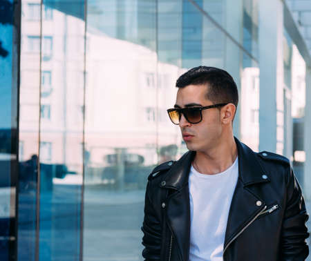 beautiful and attractive guy is standing near the glass building in the city, posing. dressed in a black leather kurta, white T-shirt, jeans and white shoes.の写真素材