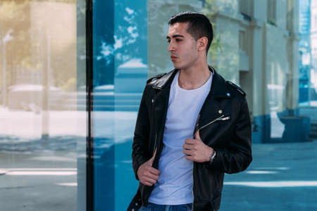 beautiful and attractive guy is standing near the glass building in the city, posing. dressed in a black leather kurta, white T-shirt, jeans and white shoes.の写真素材