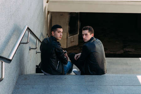 two guys in black jackets, jeans and white shoes are sitting on the steps. on the hands of a watch. socializing, meeting friends. in the city near the railings.Rear view, look at the camera. communication with a friend. guys communicate, friendshipの写真素材