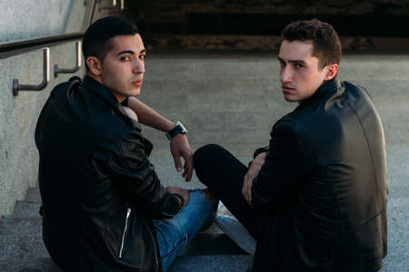 two guys in black jackets, jeans and white shoes are sitting on the steps. on the hands of a watch. socializing, meeting friends. in the city near the railings.Rear view, look at the camera. communication with a friend. guys communicate, friendshipの写真素材