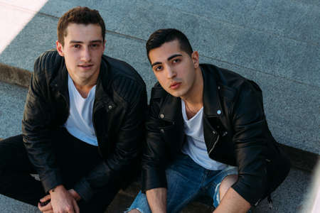 two guys in black jackets, jeans and white shoes are sitting on the steps. on the hands of a watch. socializing, meeting friends. in the city .look at the cameraの写真素材