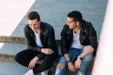 two guys in black jackets, jeans and white shoes, in white T-shirts are sitting on the steps. on the hands of a watch. socializing, meeting friends. in the city communionの写真素材