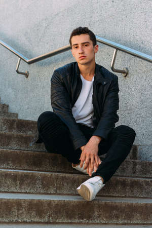 Sexy man posing sits on the steps near railing. an attractive young man In a black leather jacket, a...