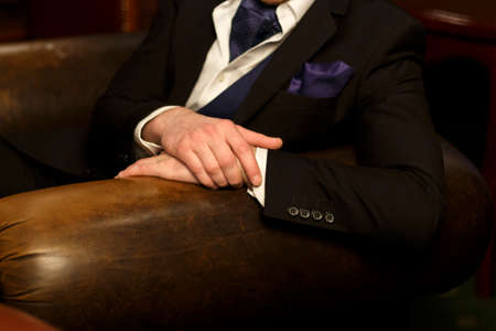 hands of the man lie on the back of the chair, he himself in a dark suit with a purple handkerchief,...