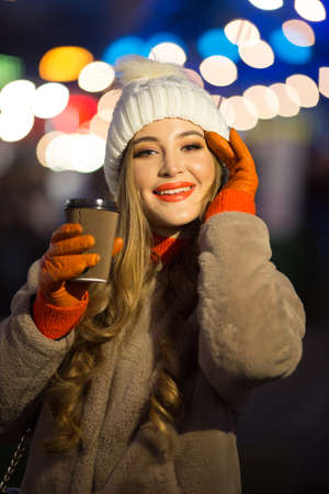 beautiful girl on the background of lights with coffee, on the street, holiday, new year. a walk through the evening city smilesの写真素材