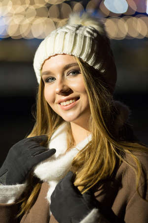 beautiful girl, in the evening, against the background of lights, a white hat and a beige coat. smiling at camera, close-up portraitの写真素材