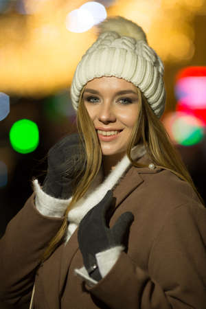 beautiful girl, in the evening, against the background of lights, a white hat and a beige coat. smiling at camera, close-up portraitの写真素材