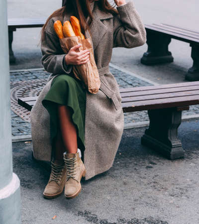 a girl in a coat and hat holds a bag with baguettes, bread, a loaf, on the street. a shopping trip, morning. sits on a benchの写真素材