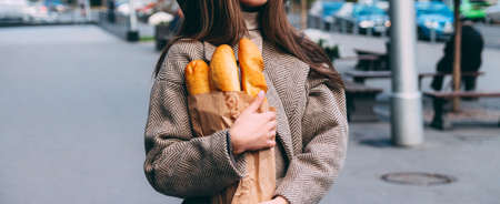 a girl in a coat and hat holds a bag with baguettes, bread, a loaf, on the street. a shopping trip, morning.の写真素材