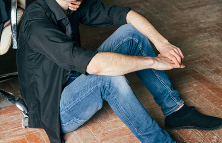 a man sits on a wooden floor, his arms around his neck. a man is sitting next to a chairの写真素材