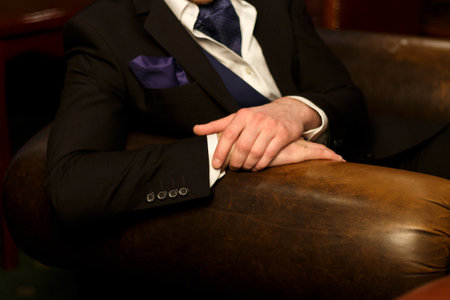 hands of the man lie on the back of the chair, he himself in a dark suit with a purple handkerchief, in a vest sits on a chairの写真素材