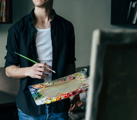 the artist holds a palette with paints and a brush and is going to paint on canvas. a man in jeans and a black shirt. hobby.の写真素材