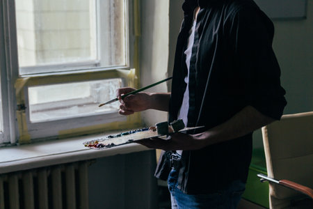 the artist holds a palette with paints and a brush and is going to paint on canvas. a man in jeans and a black shirt. hobby. across from the windowの写真素材