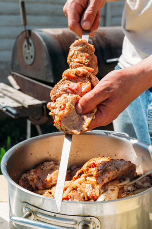 a man strings pieces of raw meat on a metal skewer to cook it on fire. Grilled shish kebab. meat on skewers lies on a white tray. barbecue. cook meat with onions and peppers.の写真素材