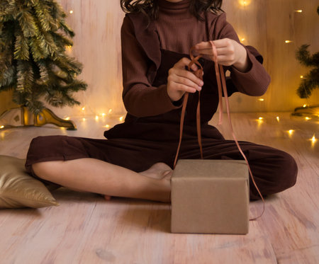 a girl in a brown dress is sitting barefoot on the floor and wrapping a gift, tying a ribbon on the boxの写真素材