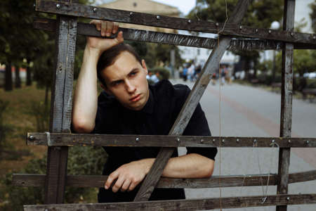 attractive guy on the street, in black, leaning on a hedge, fence