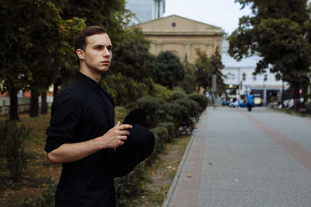 attractive guy in a hat on the street, in black, frowns and holds his hat