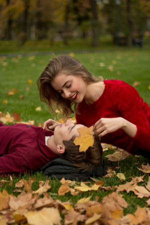 the girl and the guy in red sweaters and jeans sit in the park on the green grass in the yellow maple leaves, the girl smiles.a date in the fall.Leaves a leaf against the face of a man, protection,の写真素材