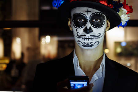 a man with a painted face of a skeleton, a dead zombie, in the city during the day. day of all souls, day of the dead, halloween, ghost walk in a hatの写真素材