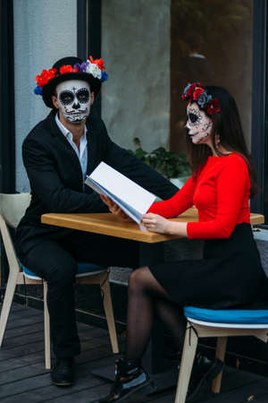 A couple, wearing skull make-up for. All souls day. Boy and girl sugar skull makeup.painted for halloween sit in a cafe. dead in the city. zombie walk.day of the dead holiday in mexicoの写真素材