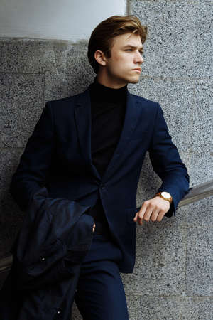 portrait of an attractive young man in a blue suit in the city on gray background.watch on hand, gold wrist watch.pensiveの写真素材