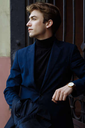 portrait of an attractive young man in a blue suit in the city on gray background.watch on hand, gold wrist watch.pensiveの写真素材