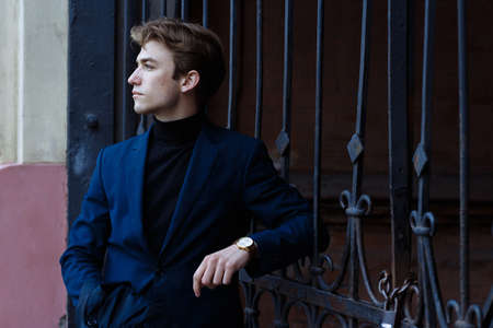 portrait of an attractive young man in a blue suit in the city on gray background.watch on hand, gold wrist watch.pensiveの写真素材