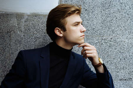 portrait of an attractive young man in a blue suit in the city on gray background.watch on hand, gold wrist watch.pensiveの写真素材